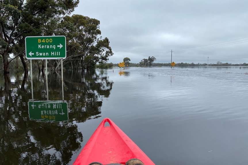 A red canoe paddles down a road with a road sign on the left on a cloudy day. It is flooded
