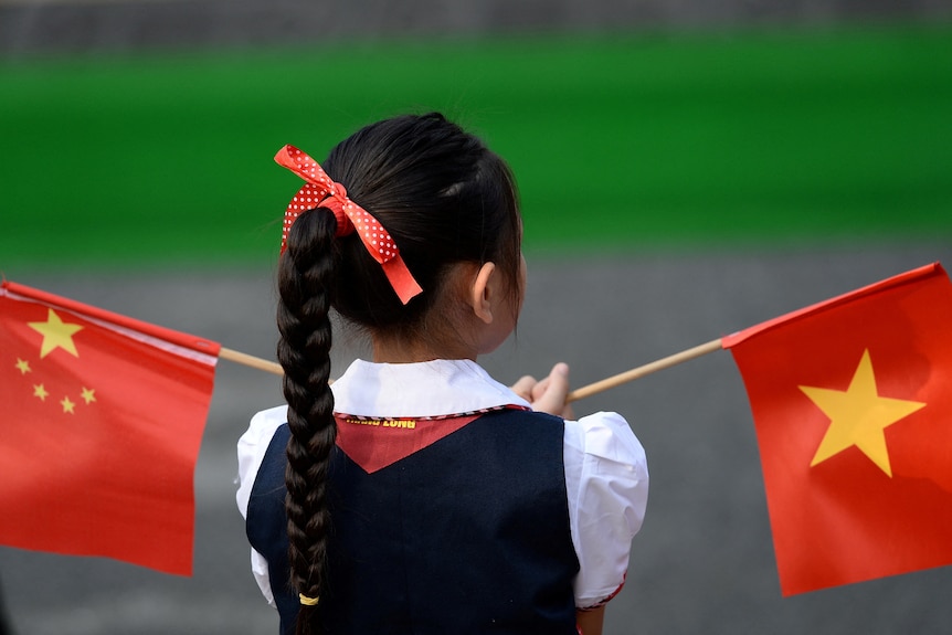Back of girl with long black plait and red ribbon holding a Vietnamese and a Chinese flag in each hand.