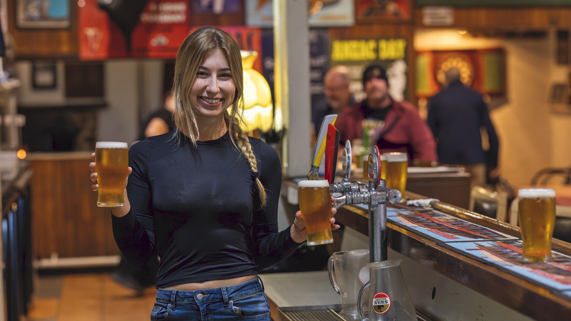 A young woman with blond hair smiles and holds a pint of beer in each hand