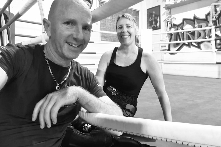 A man leans on the ropes of a martial arts ring, a woman sits on the mat, they are both smiling at the camera.