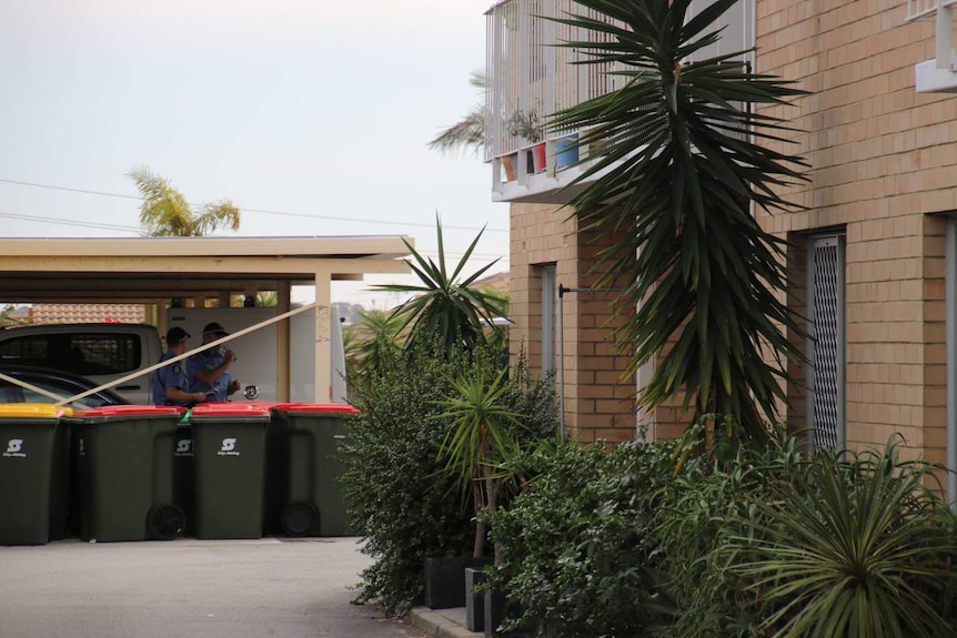 Two police officers stand under a carport outside a block of units.