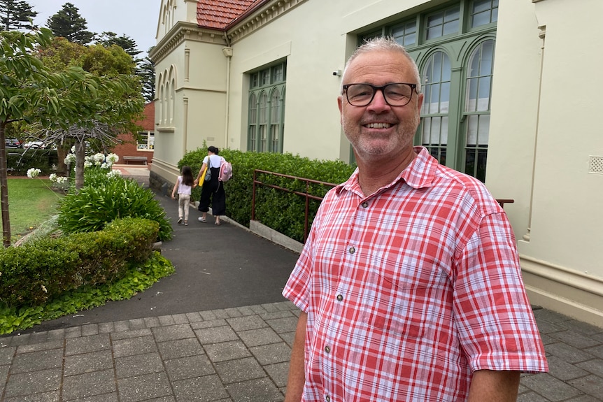 A smiling man wearing a red checked short sleeve shirt in front of primary school