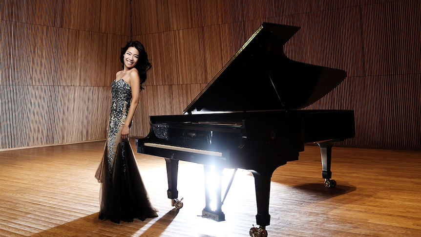 A woman stands on stage in front of a grand piano. She wears a sequined gown.