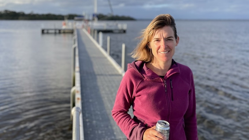 Megan Holbeck smiles while standing on a windy pier holding a drink can. She has blonde hair and wears a purple fleece top.