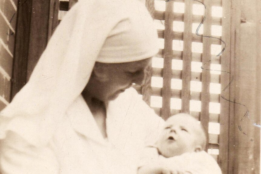 A black and white photograph of an older woman in a nursing uniform holding a baby outside.