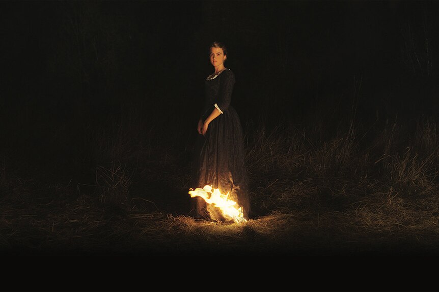 A woman with neutral expression in 18th century period dress stands alone in field at night with bottom of dress on fire.