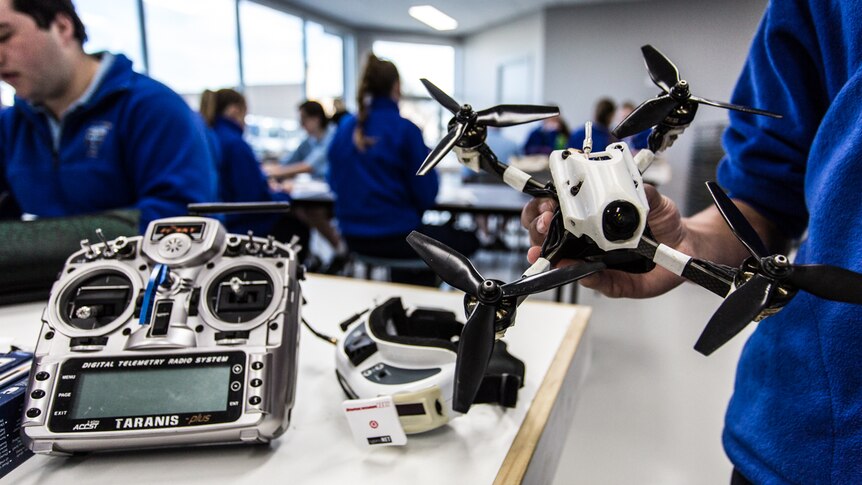 A close up of a drone held in a hand with a back drop of a classroom.