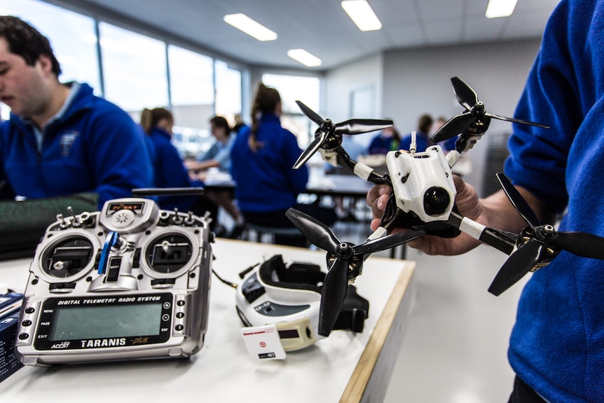 A close up of a drone held in a hand with a back drop of a classroom.