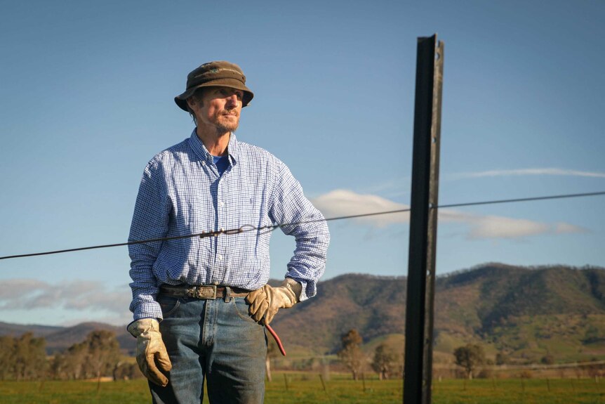 Tony Jarvis wears a hat, gloves, jeans and blue shirt, he's standing behind a fence he's rebuilding, with green paddocks behind.