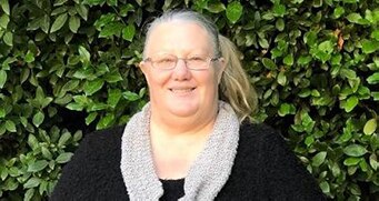 In front of a green hedge, a woman in glasses and black jumper, with light scarf, smiles.