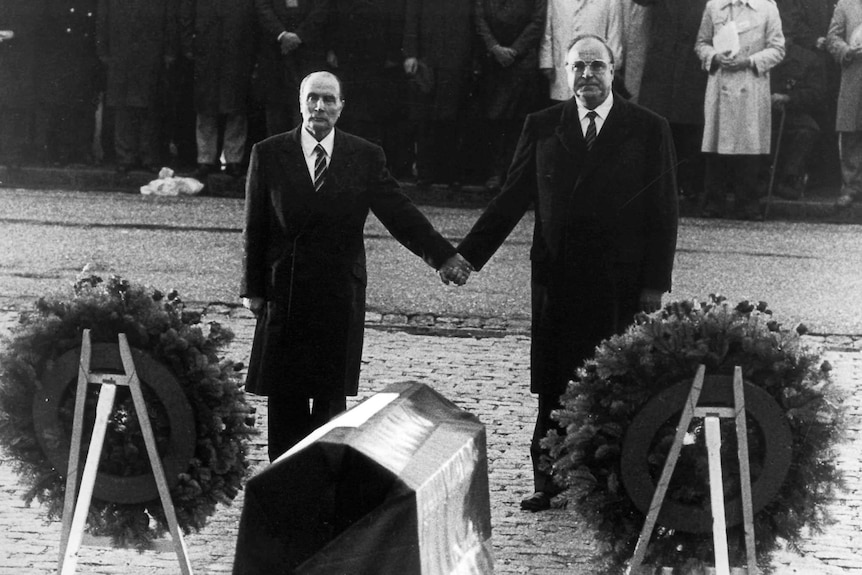 Francois Mitterrand and Helmut Kohl stand holding hands at a ceremony.