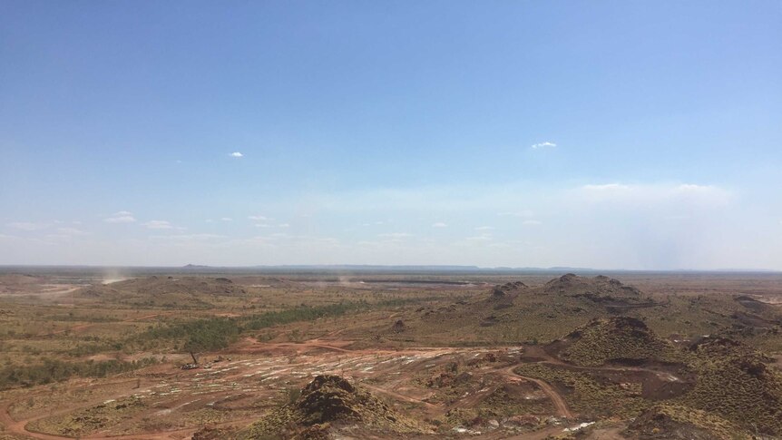 The Pilbara landscape of rolling hills and a mine under construction sits beneath a blue sky.