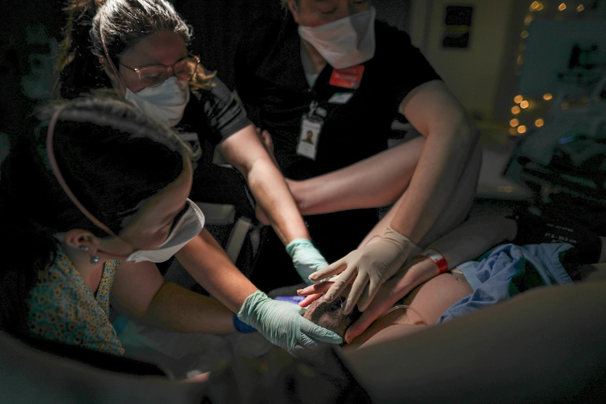 Three midwives reach towards a woman who is giving birth on a hospital bed.