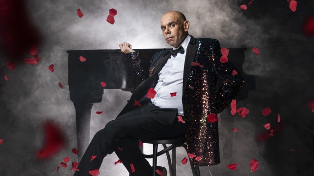 A man in a black sequin blazer and bowtie leans casually back in a chair. There is a piano in the background.