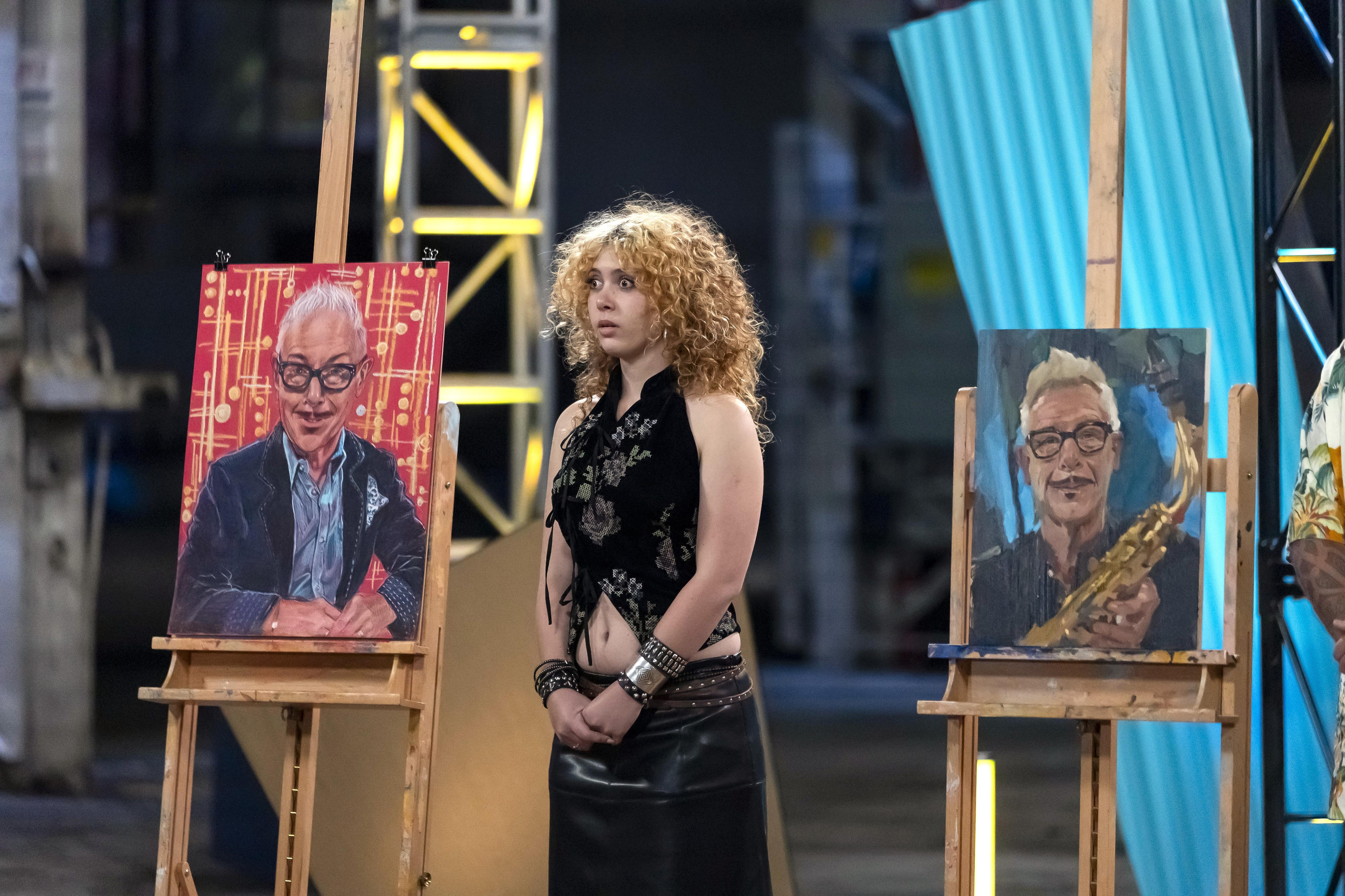 A young woman with curly hair stands in wide-eyed shock on a TV set with a portrait of Kirk Pengilly behind her.