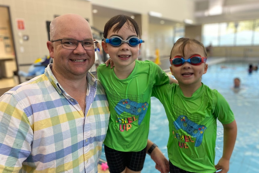 Will and Henry are wet from the pool and wearing goggles, standing with their dad.