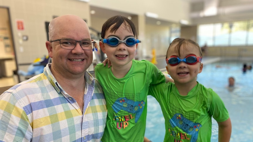 Will and Henry are wet from the pool and wearing goggles, standing with their dad.