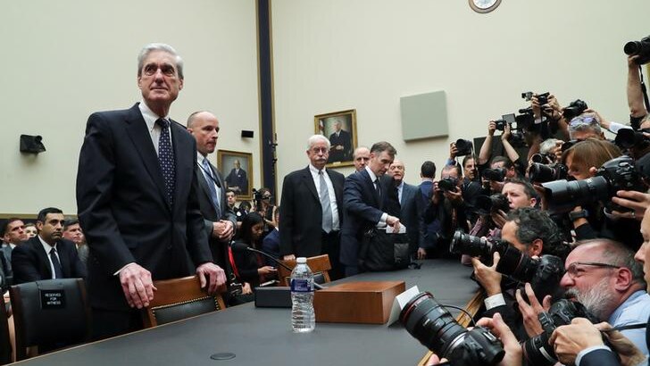 Robert Mueller looks to the left as he stands at a table with his hands on a chair as photographers take pictures of him.