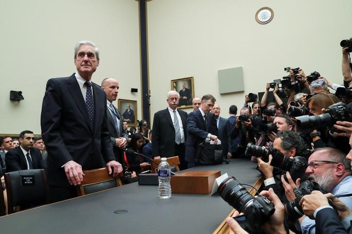 Robert Mueller looks to the left as he stands at a table with his hands on a chair as photographers take pictures of him.