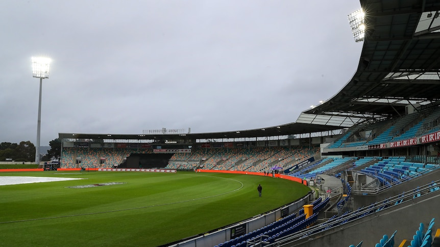 A picture of a cricket ground mostly empty aside from a handful of people, with covers on the pitch in the background. 