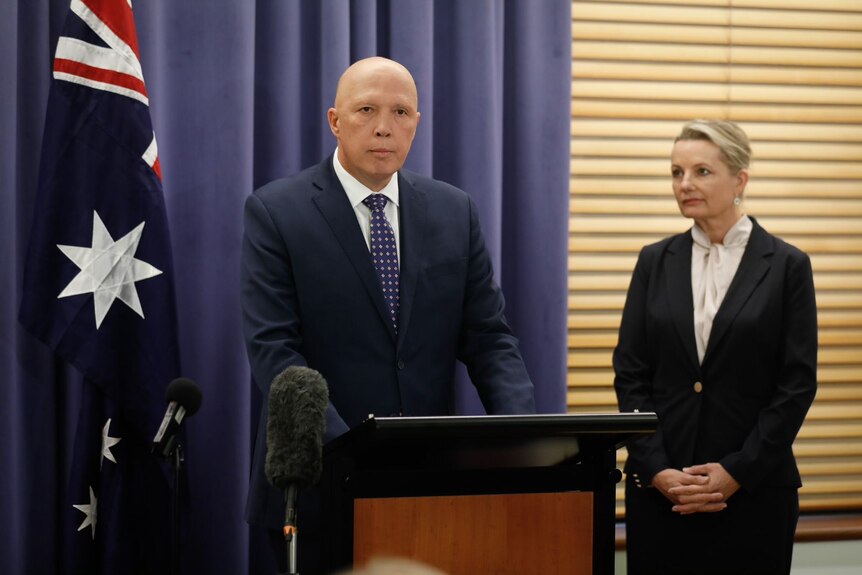 Peter Dutton stands at a lectern with his lips pursed. Sussan Ley stands to his right, watching