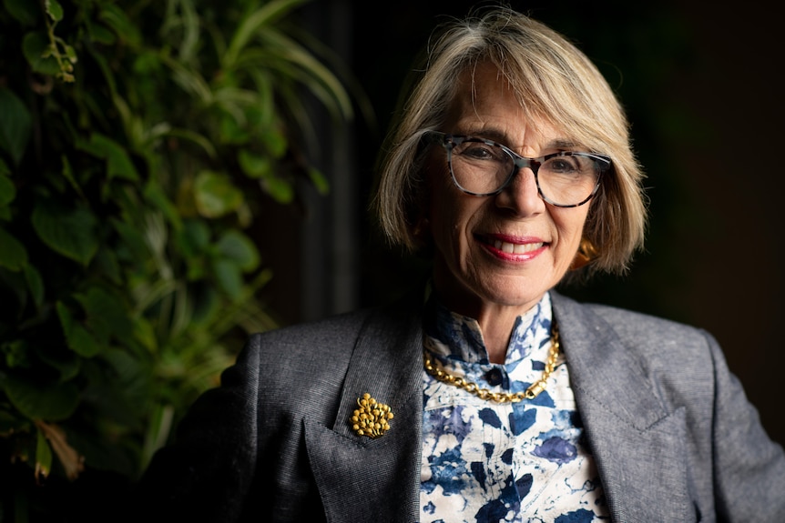 A smiling woman with glasses smiles with greenery on the left