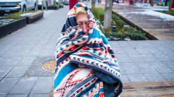 A woman sits on the street wrapped in a blanket.