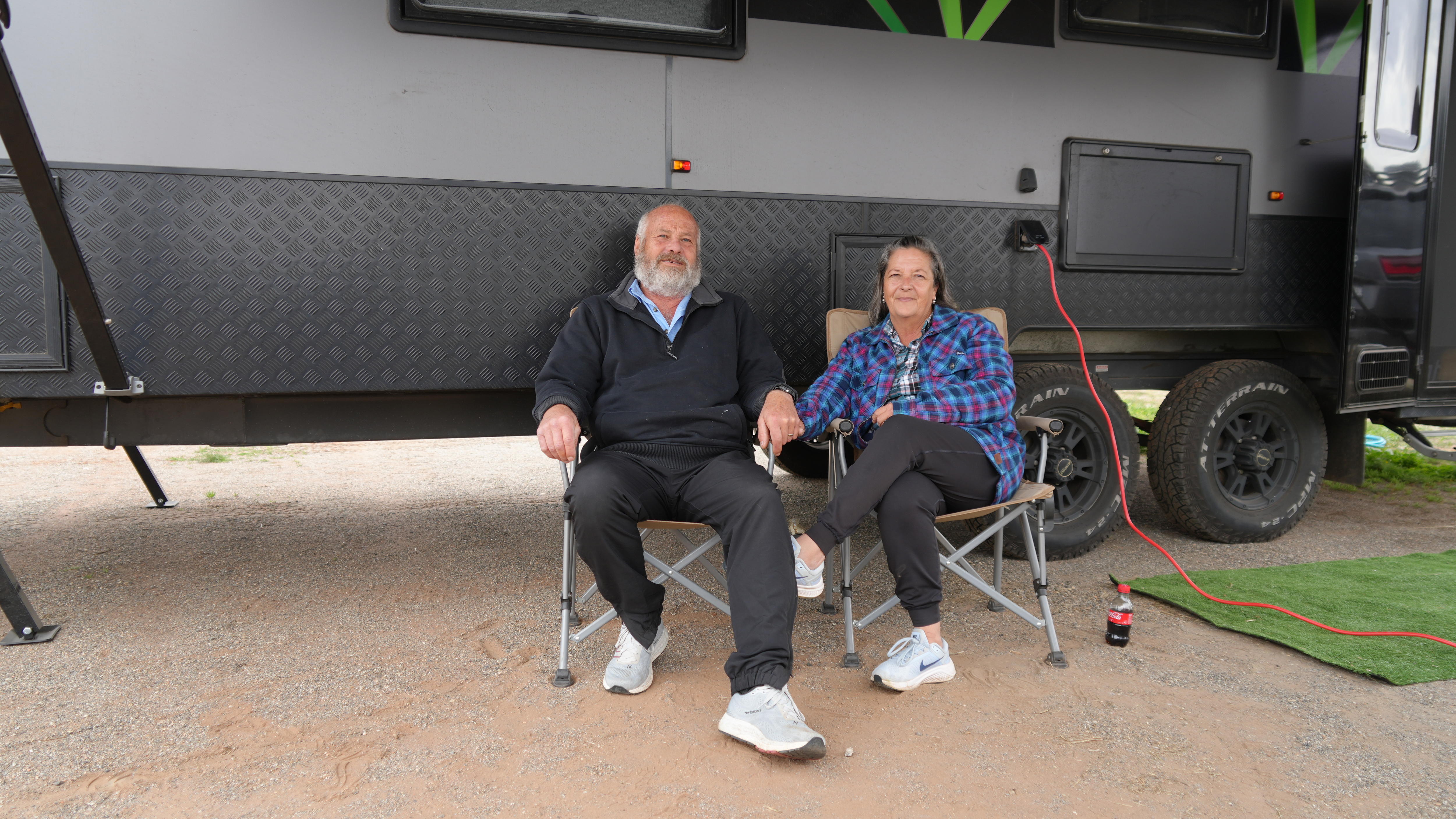 Couple sits on fold up chairs in front of caravan.