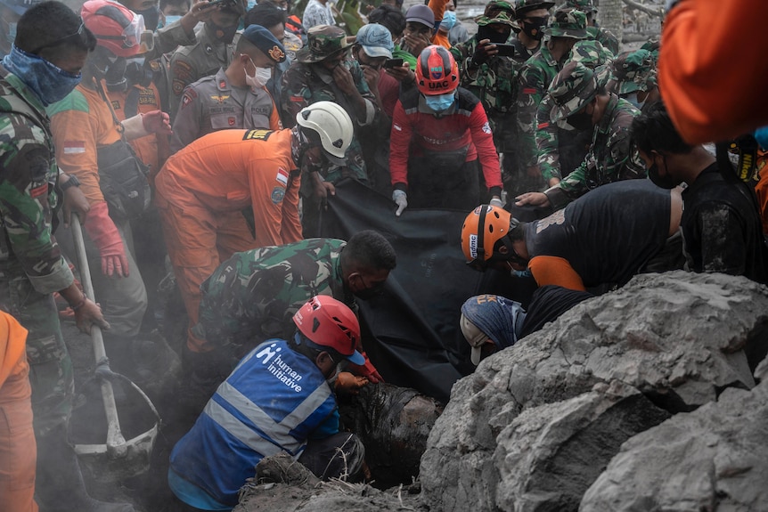 Rescuers in dusty high vis and military gear work to free a body trapped under a grey slab of volcanic ash.
