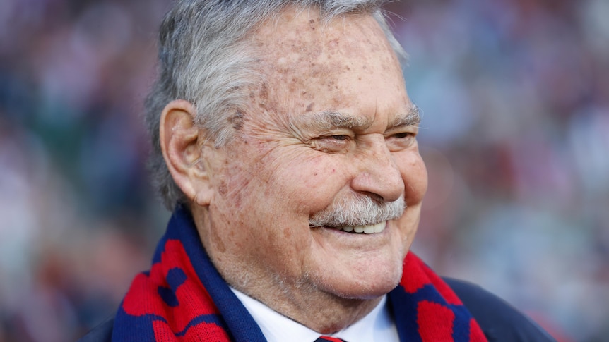 An older man wearing a red and blue AFL scarf, in a suit, smiles widely in happiness.