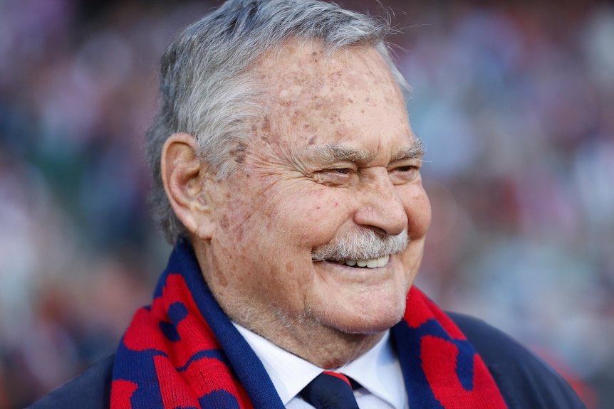 An older man wearing a red and blue AFL scarf, in a suit, smiles widely in happiness.