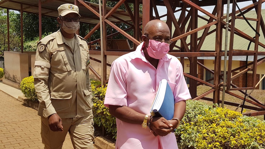 Wearing a pink shirt and shorts, Paul Rusesabagina is escorted in handcuffs by armed guards.