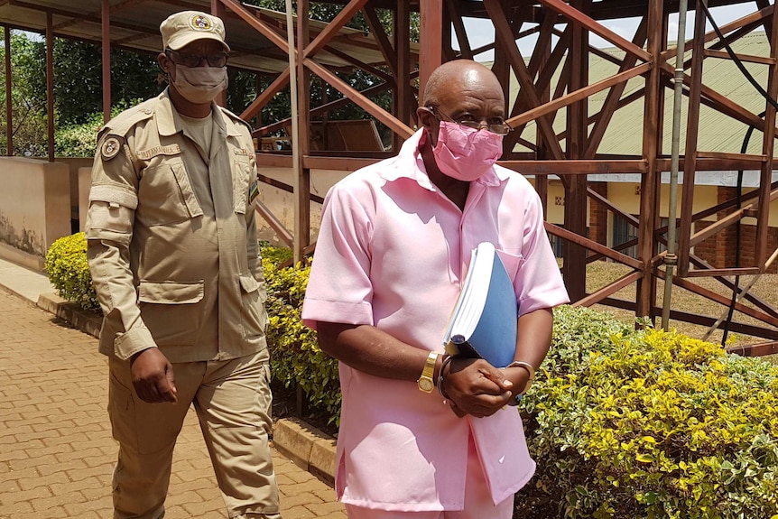 Wearing a pink shirt and shorts, Paul Rusesabagina is escorted in handcuffs by armed guards.