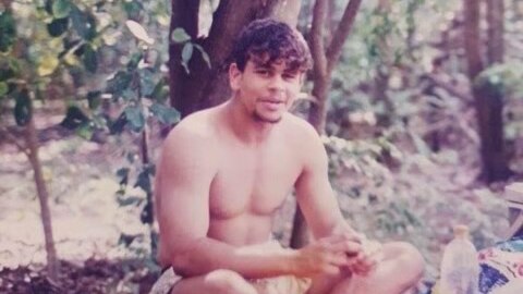 An old colour photo of a young man sitting in bushland with no shirt on