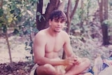 An old colour photo of a young man sitting in bushland with no shirt on
