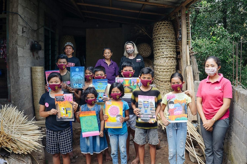 A group of Balinese children hold up paintings as other adults watch. Forest is behind them