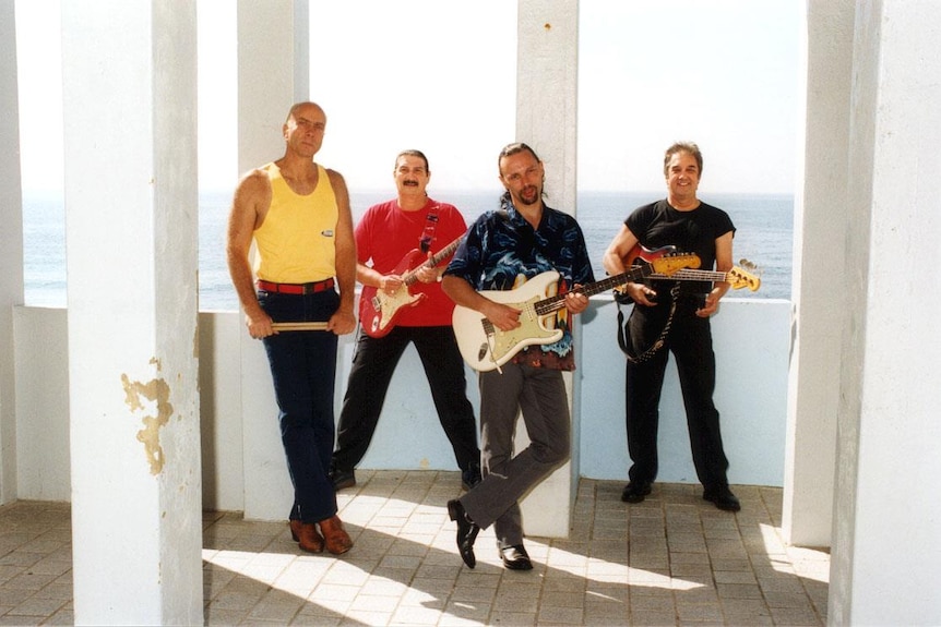 A photo of The Atlantics band, four men posing with guitars on a wooden deck in front of the sea