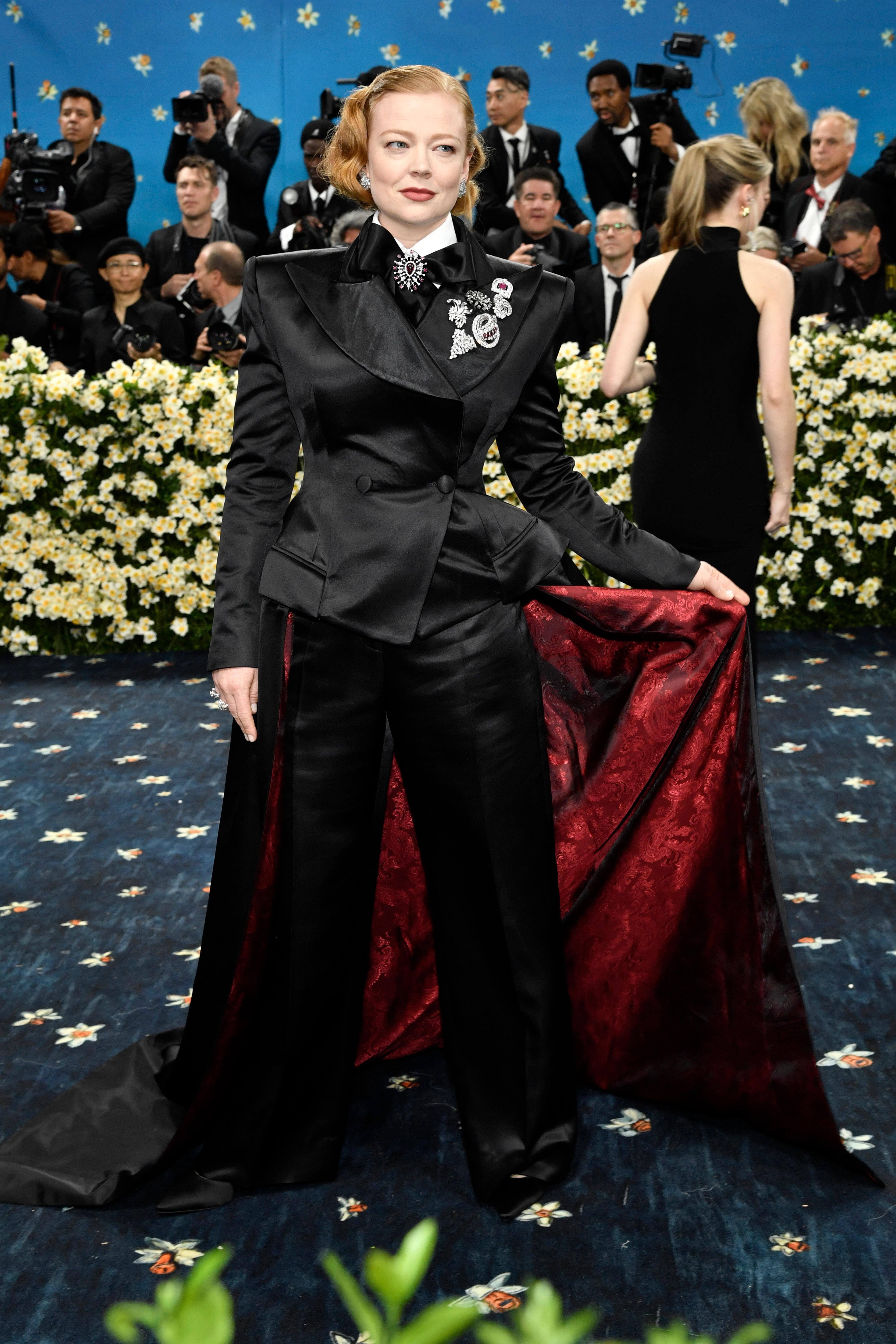 Sarah Snook wearing a black suit with a long, red-lined cape
