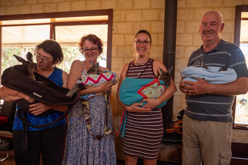 Four people stand smiling at the camera, holding blankets with joeys and possums inside