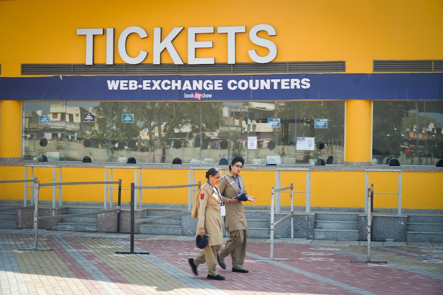 Two women walk past a sign saying TICKETS in front of a bright yellow wall