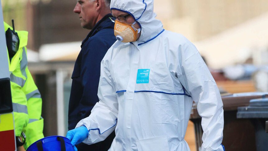 A man in a full white suit carries plastic bags and a blue box from a Manchester property.