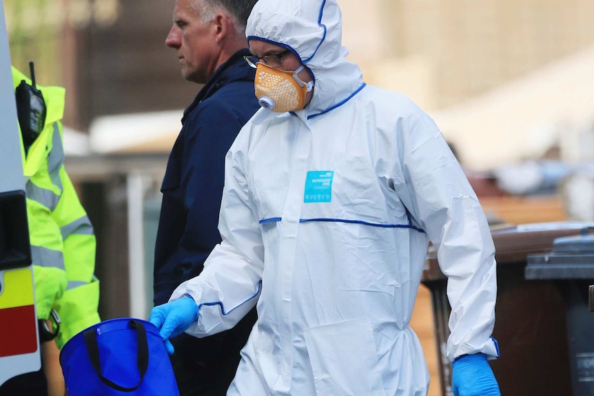 A man in a full white suit carries plastic bags and a blue box from a Manchester property.