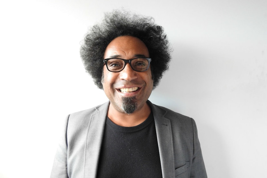 A smiling man with curly black hair and black glasses wearing a suit stands in front of a plain white wall.