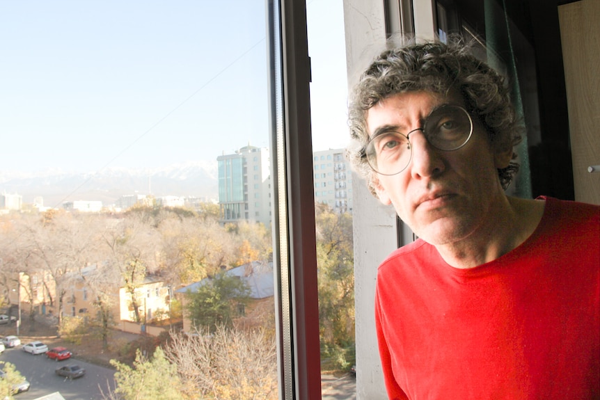 A close up of a man with grey hair wearing glasses and a red tshirt standing next to a window.