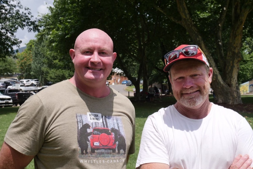 A tall bald man and another man in a red cap smile side by side. Trees line the background.