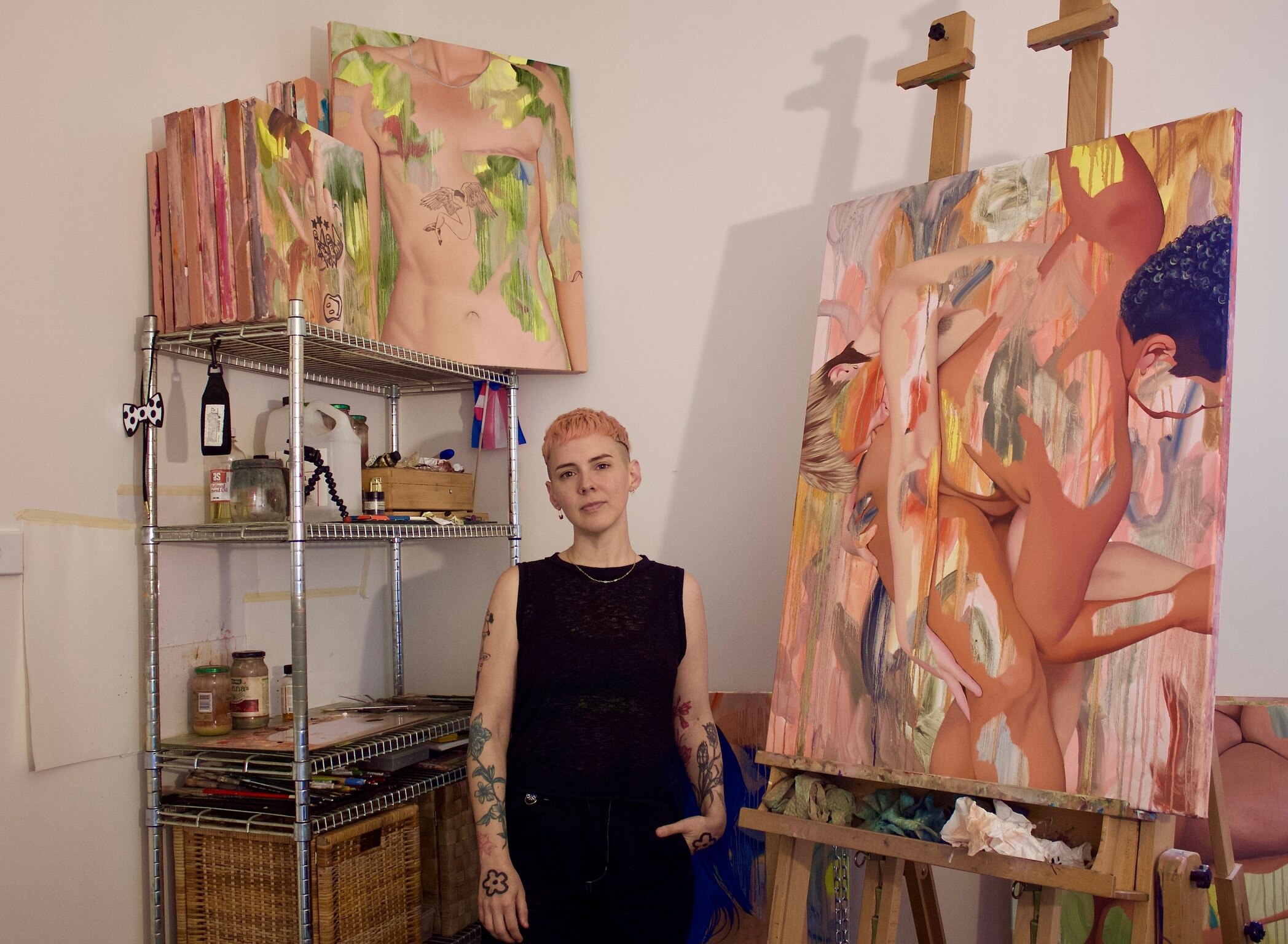 Artist Kim Leutwyler in dark singlet with short blonde hair standing in an art studio near paintings and shelves of art supplies