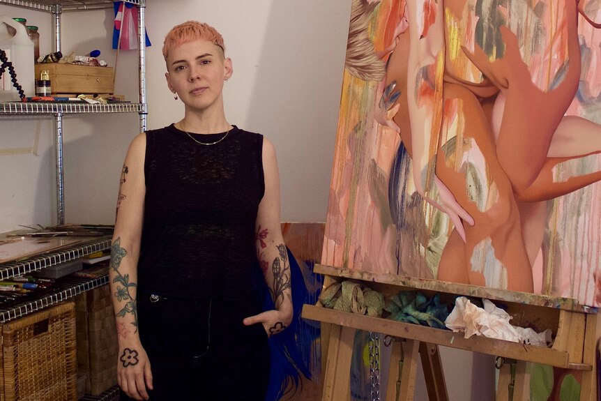 A white person in a dark singlet with short blonde hair, standing in an art studio near paintings and shelves of art supplies