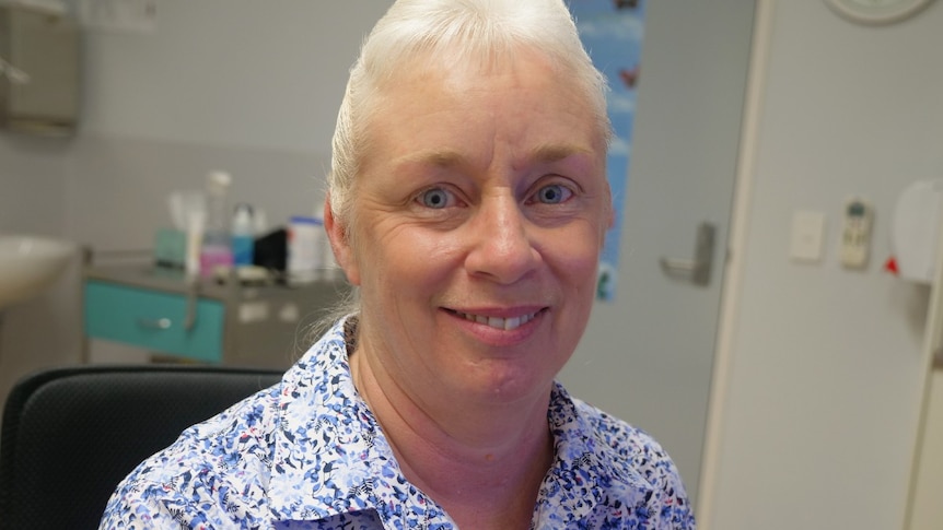 Adele Love, blue eyes, smiles at the camera, blue and white patterned shirt, doctor's clinic behind.