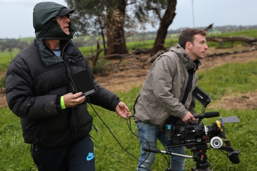 Man in black raincoat with another man holding camera