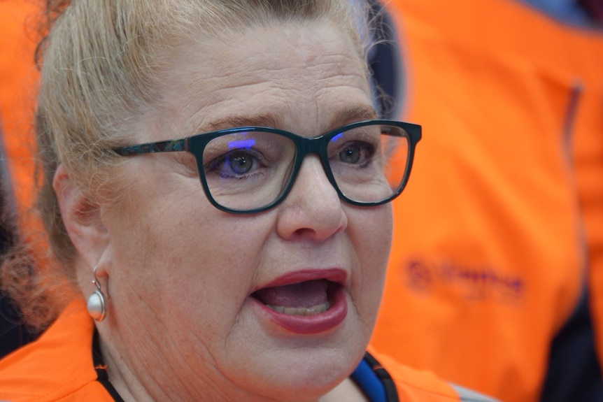 A close up shot of a woman wearing glasses standing in front of people wearing orange hi-vis